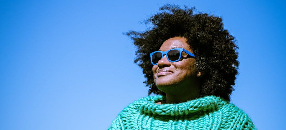 Image of Aja posing on a clear day, wearing a thick knit green jumper and blue-framed sunglasses