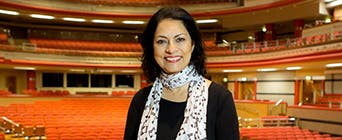 A headshot photo of Anita Bhalla standing onstage at a theatre with seats in the background. 