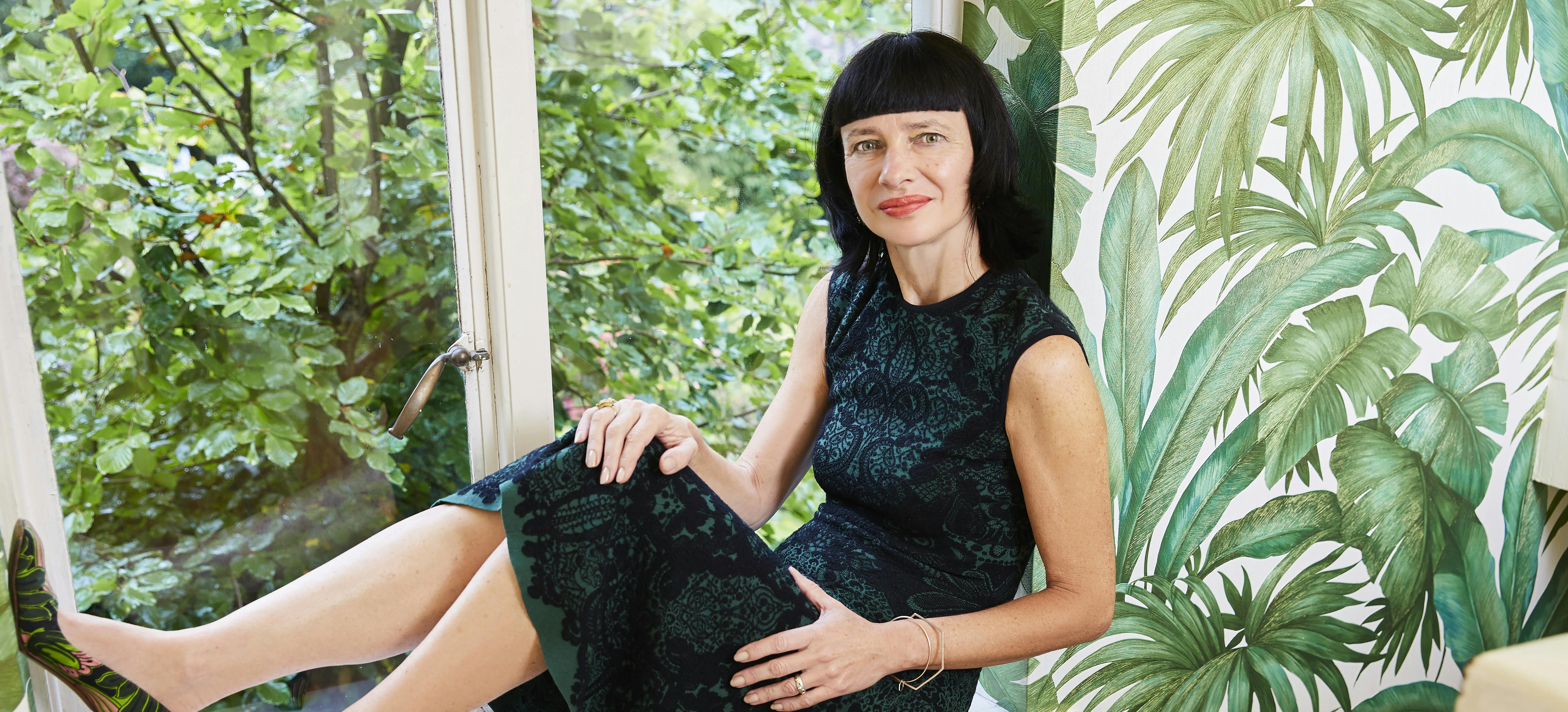 Image shows Carry sat in a window wearing a green dress and green leaf print wallpaper behind her