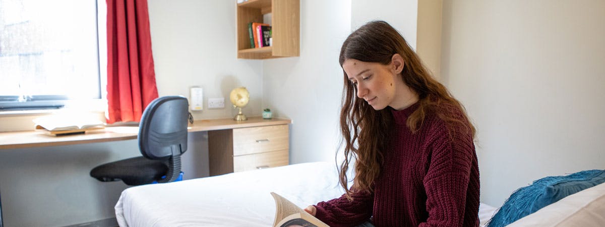 Student sits on her bed reading in her bedroom in university accommodation