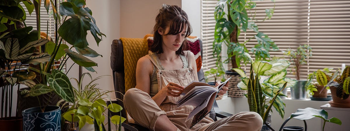 Student sat on a chair in her home reading, surrounded by houseplants.