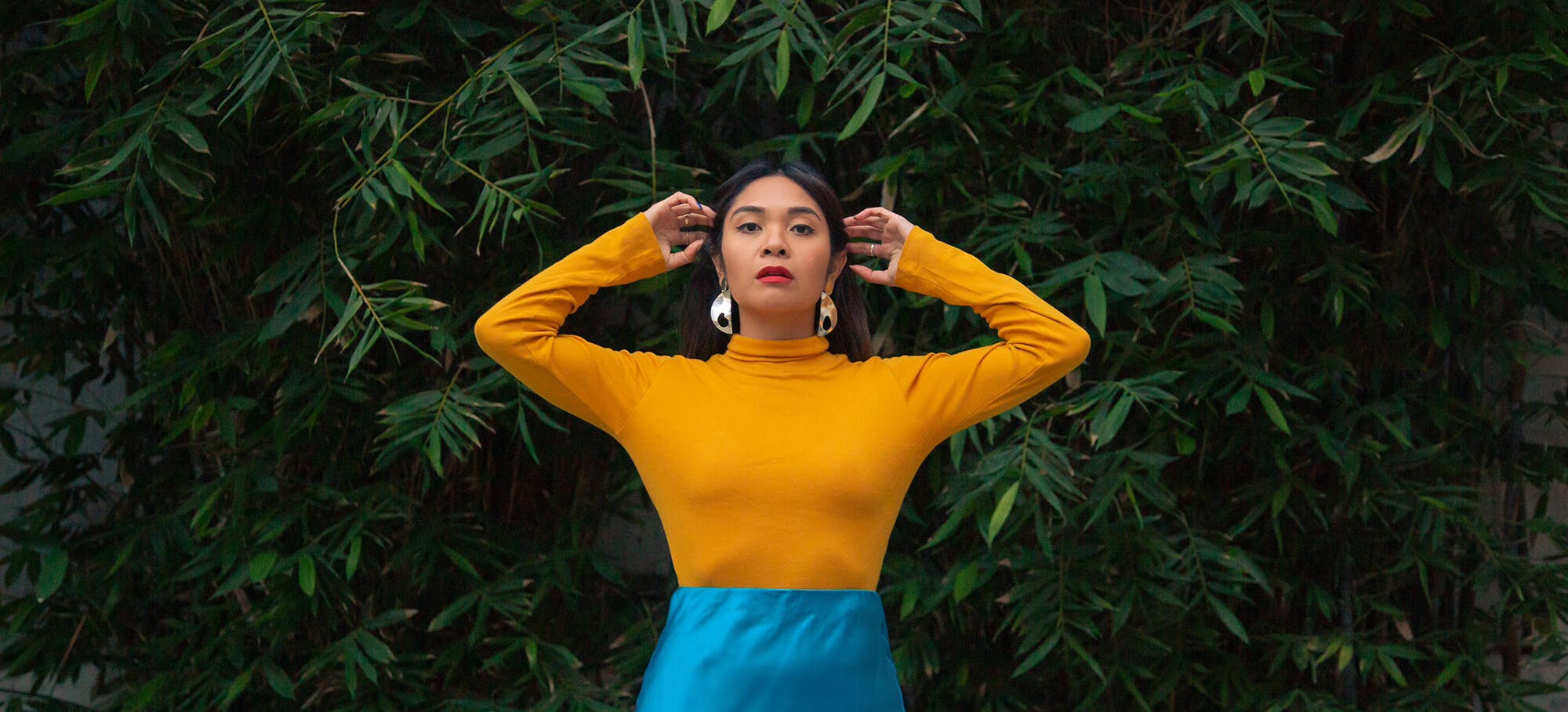 Image of Kara outdoors with her arms raised in front of foliage, wearing a bright yellow top and blue skirt