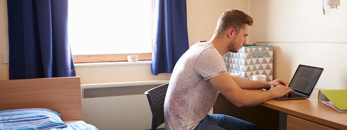 A male student sits in his accommodation on his laptop. 